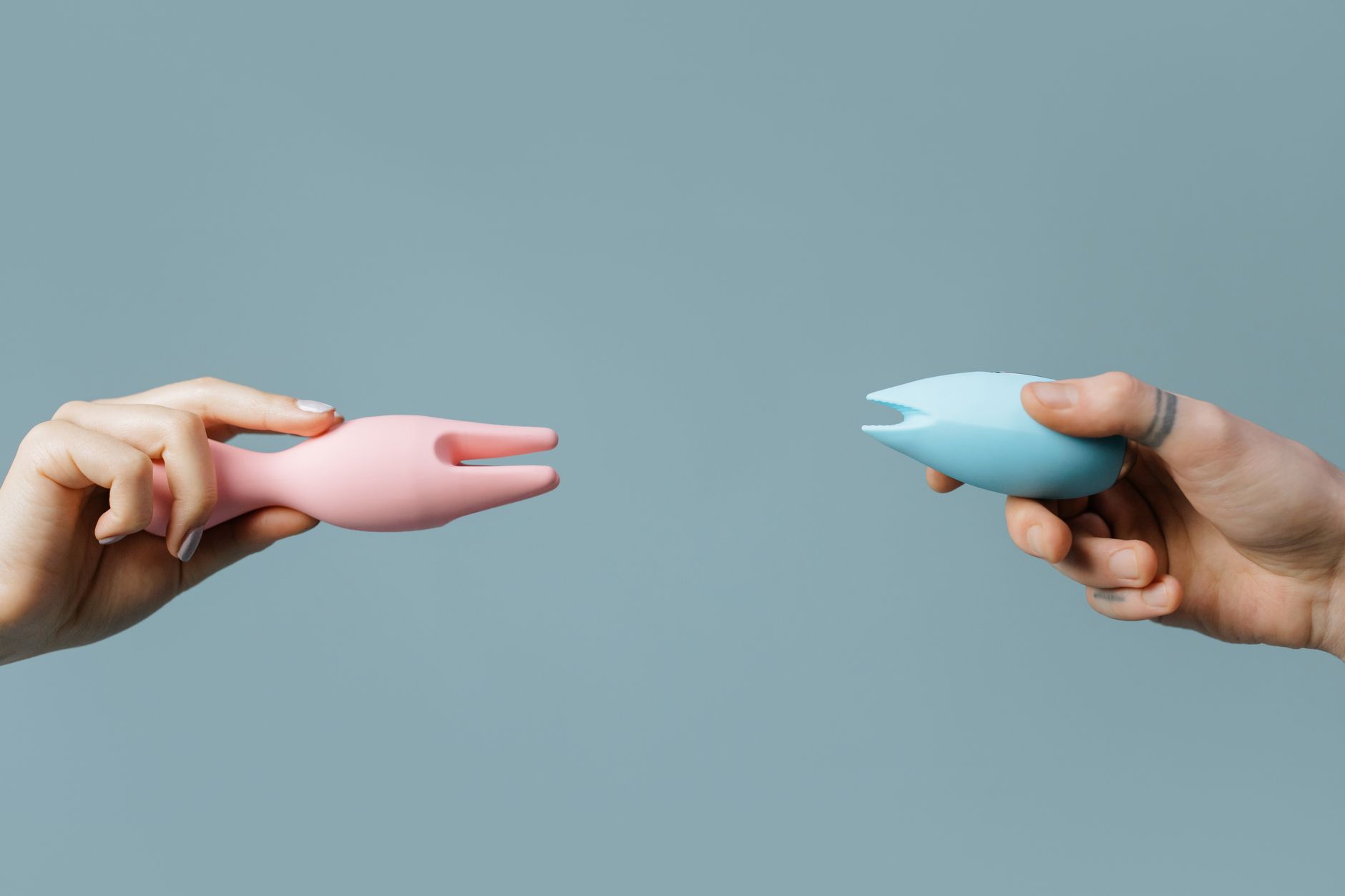Two hands holding pink and blue silicone vibrators against a pastel background, symbolizing choice and personal pleasure.